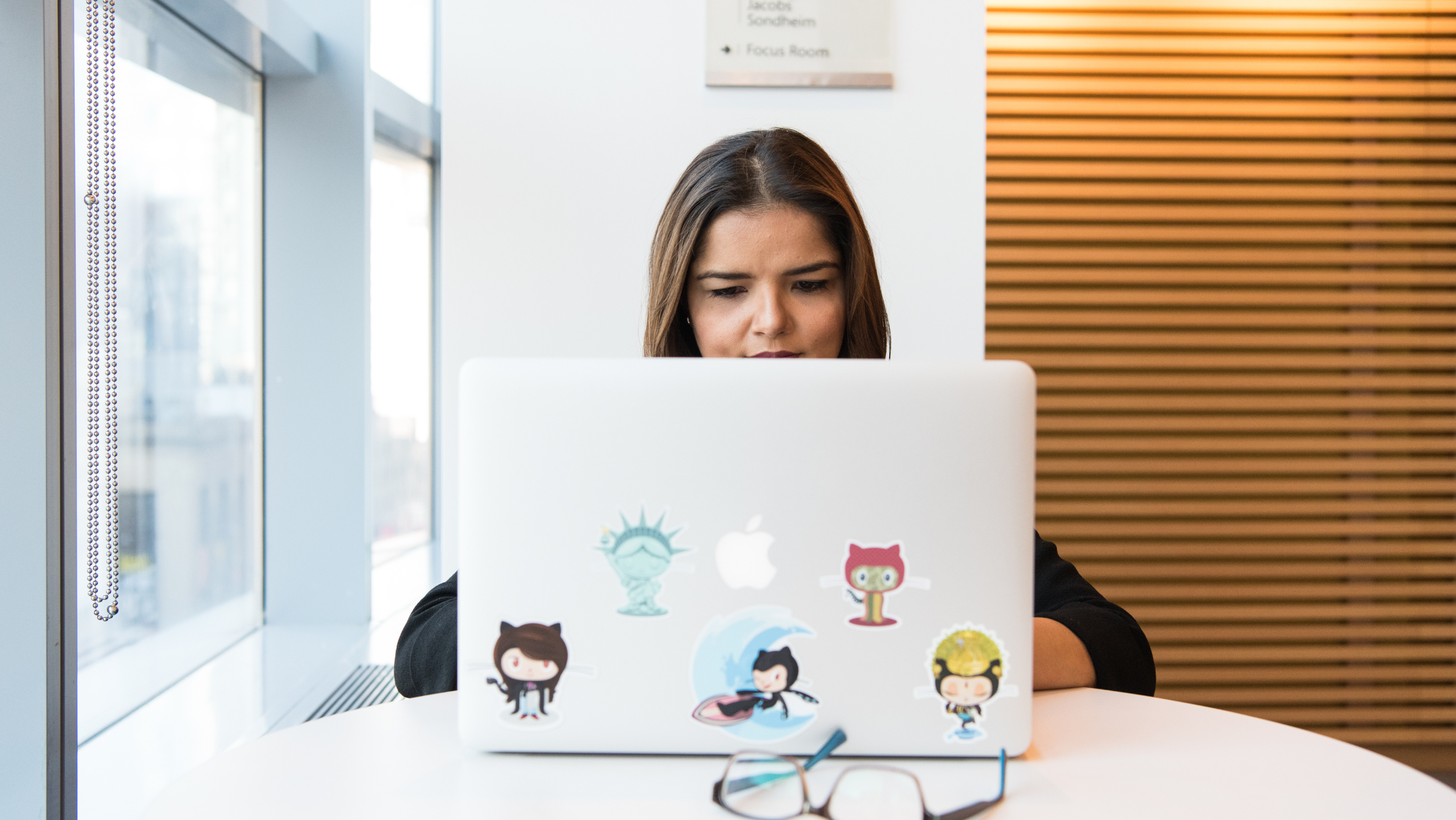 Pessoa usando um notebook adesivado com logos do GitHub, trabalhando em um ambiente de escritório com grandes janelas ao fundo.